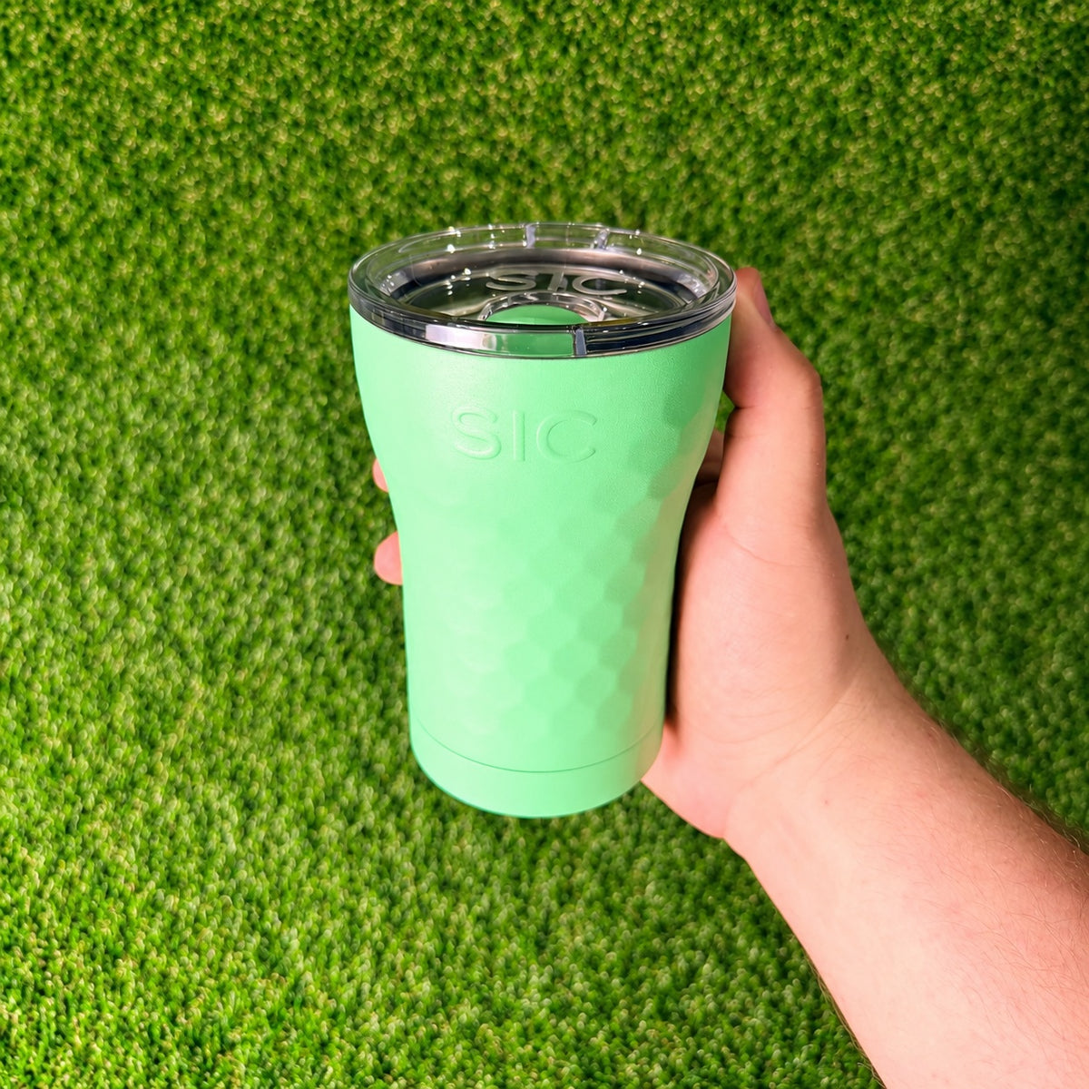 Hand holding a SIC Hammered Mint 12 oz. tumbler over green turf